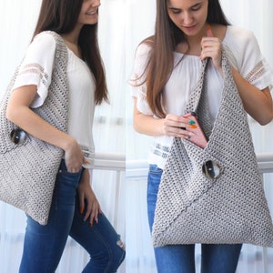 Op de afbeelding: Een grijze gehaakte tote bag met een grote knoopsluiting. De tas wordt over de schouder gedragen door iemand die een wit shirt en jeans draagt.