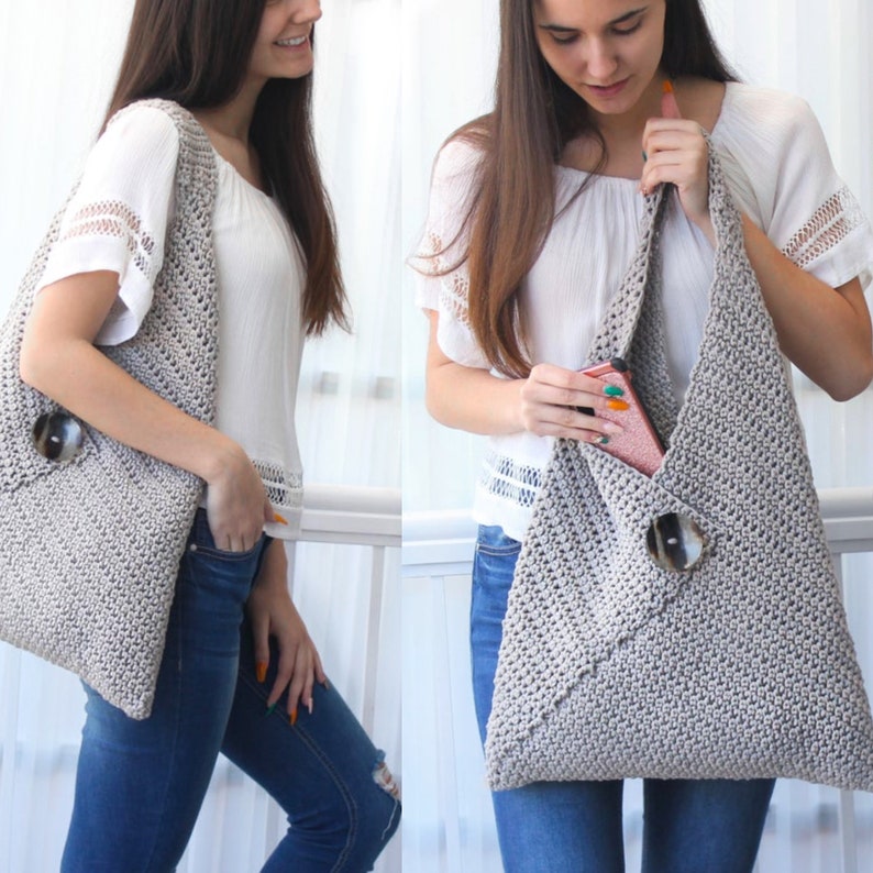 Op de afbeelding: Een grijze gehaakte tote bag met een grote knoopsluiting. De tas wordt over de schouder gedragen door iemand die een wit shirt en jeans draagt.
