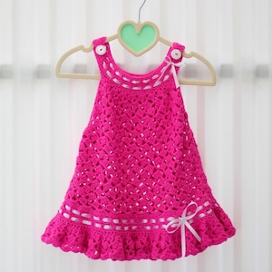 May include: A pink crocheted baby dress with white ribbon trim and white buttons on the straps.