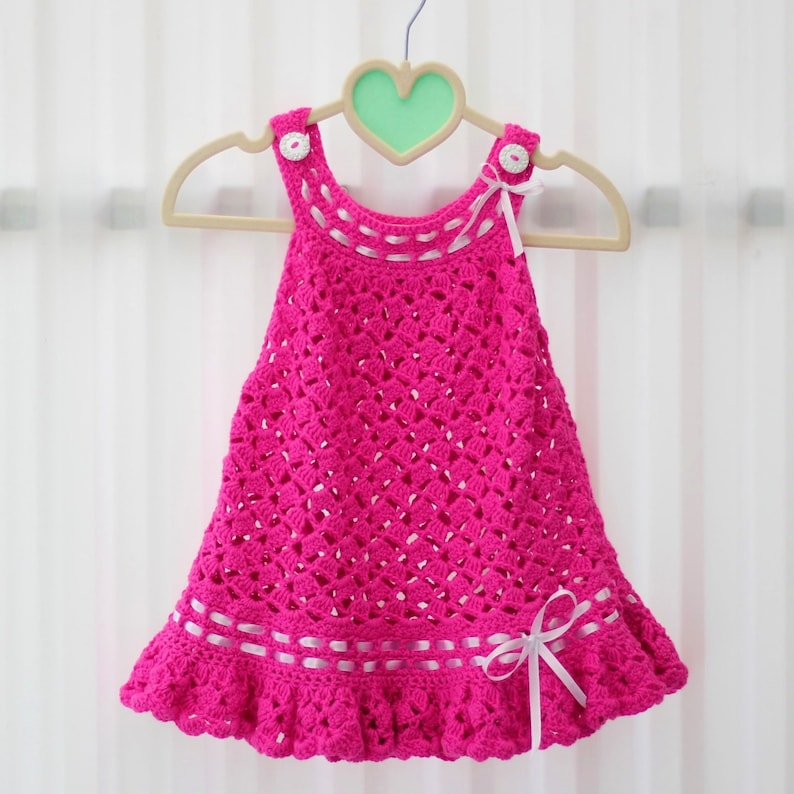 May include: A pink crocheted baby dress with white ribbon trim and white buttons on the straps.
