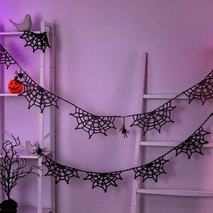 May include: A black crocheted spider web garland featuring multiple web segments and small attached black spiders. The garland is draped across two white ladder-style shelves, creating a decorative display with a dark, intricate pattern.