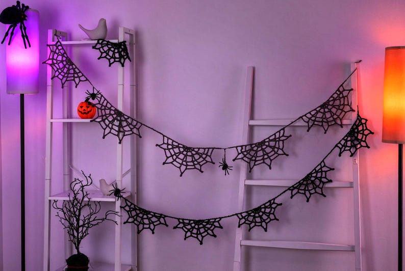 May include: A black crocheted spider web garland featuring multiple web segments and small attached black spiders. The garland is draped across two white ladder-style shelves, creating a decorative display with a dark, intricate pattern.