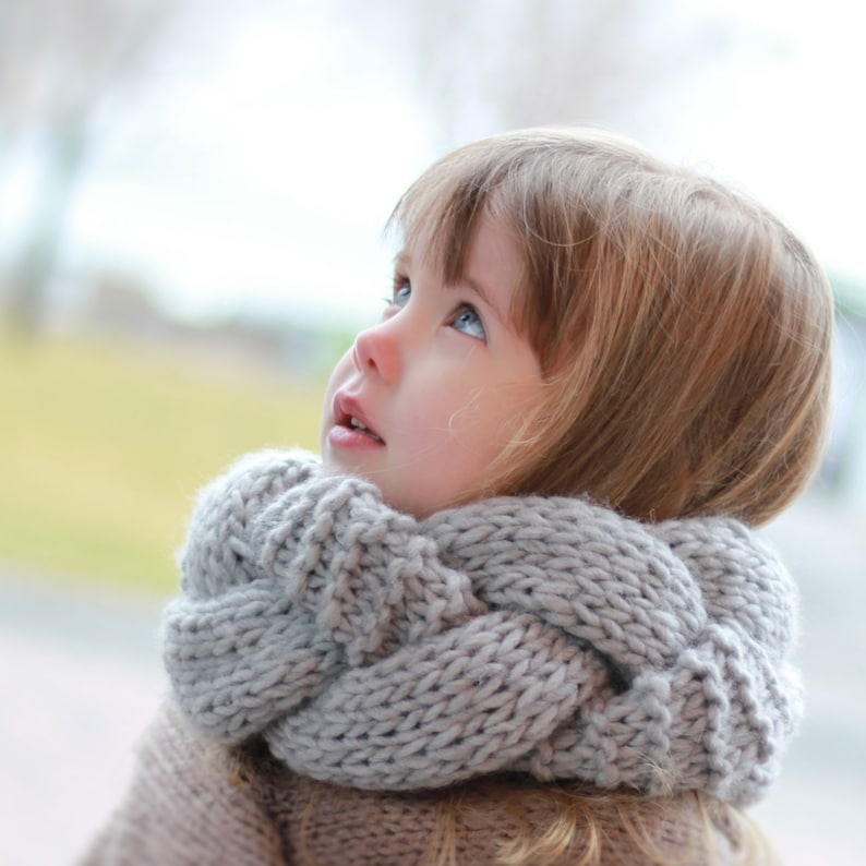 May include: A young girl with blonde hair wearing a gray knitted scarf looks up at the sky.