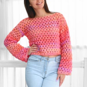 May include: A crocheted sweater in shades of pink and orange. The open weave design features long sleeves and a boat neck. The sweater is a cropped style, perfect for layering or wearing on its own. The yarn colors transition from light to dark.
