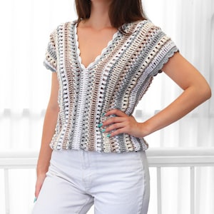 Puede incluir: Un top de ganchillo de manga corta con cuello en V en una paleta de colores neutros de blanco, gris y beige. El top presenta un escote festoneado y un dise&ntilde;o de tejido abierto texturizado. La prenda est&aacute; hecha a mano.