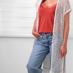 May include: A white crochet open-front vest featuring a loose net pattern, short sleeves, and long fringe trim at the hem, worn over a rust-colored camisole with lace trim and light blue denim jeans with a classic button and zipper fly closure.