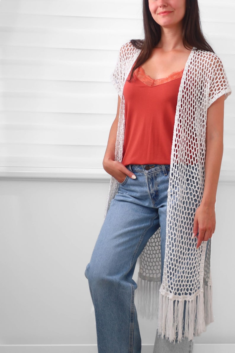 May include: A white crochet open-front vest featuring a loose net pattern, short sleeves, and long fringe trim at the hem, worn over a rust-colored camisole with lace trim and light blue denim jeans with a classic button and zipper fly closure.