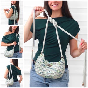 May include: A crescent-shaped crochet handbag featuring a variegated pattern of light gray, white, and soft green yarn. The bag includes a long, adjustable light gray strap with metallic end caps and a small oval brand tag on the front exterior.