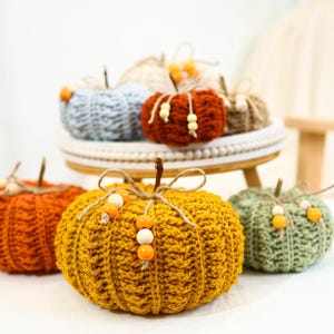May include: A collection of handmade crochet pumpkins in various colors, including orange, yellow, and green. Each pumpkin features a stem and is adorned with wooden beads. Some pumpkins are displayed on a decorative stand, creating a fall-themed display.