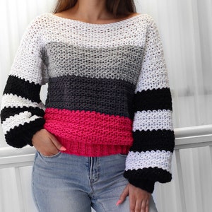May include: A striped crochet sweater with a white, gray, black, and pink color scheme. The sweater has a loose fit and long sleeves.