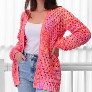 May include: A vibrant, open-front crochet cardigan in shades of pink and orange. The handmade garment features a loose, open weave design with long sleeves. The cardigan is a stylish, comfortable piece for layering.