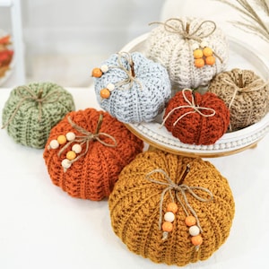 May include: A collection of handmade crochet pumpkins in various colors, including orange, green, and cream. Each pumpkin is adorned with twine and wooden bead accents, creating a rustic and autumnal aesthetic. Some pumpkins are displayed on a white tray.