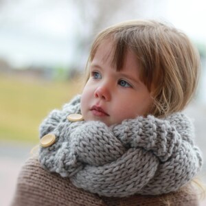 May include: A young girl wearing a gray knitted scarf with wooden buttons. The scarf is thick and warm, perfect for cold weather.