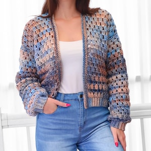 May include: A crocheted cardigan in shades of blue, brown, and tan. The open-front sweater has long sleeves and a textured, open weave design. The model is wearing blue jeans and a white top.