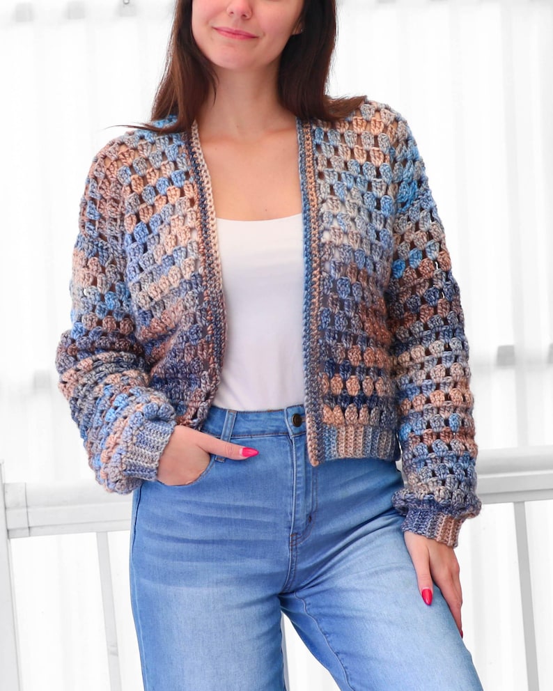 May include: A crocheted cardigan in shades of blue, brown, and tan. The open-front sweater has long sleeves and a textured, open weave design. The model is wearing blue jeans and a white top.