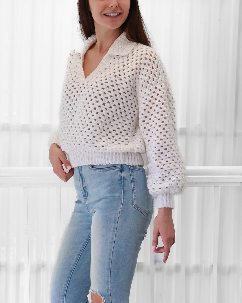 Peut inclure: Pull en crochet blanc &agrave; col et manches longues. Le pull est ample et pr&eacute;sente un motif textur&eacute;. Le mannequin porte le pull avec un jean bleu.
