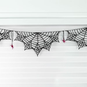 May include: A black crocheted spider web garland featuring three distinct web sections connected by a string. Small, bright pink crocheted spiders are attached to the web segments and the connecting string, creating a detailed hanging decoration.