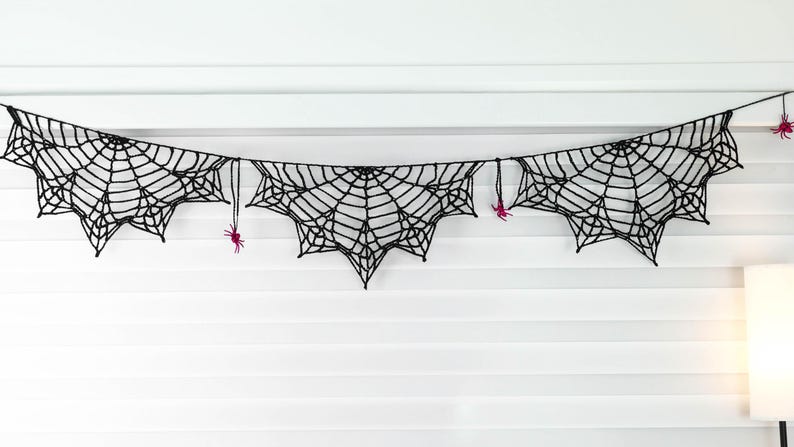 May include: A black crocheted spider web garland featuring three distinct web sections connected by a string. Small, bright pink crocheted spiders are attached to the web segments and the connecting string, creating a detailed hanging decoration.