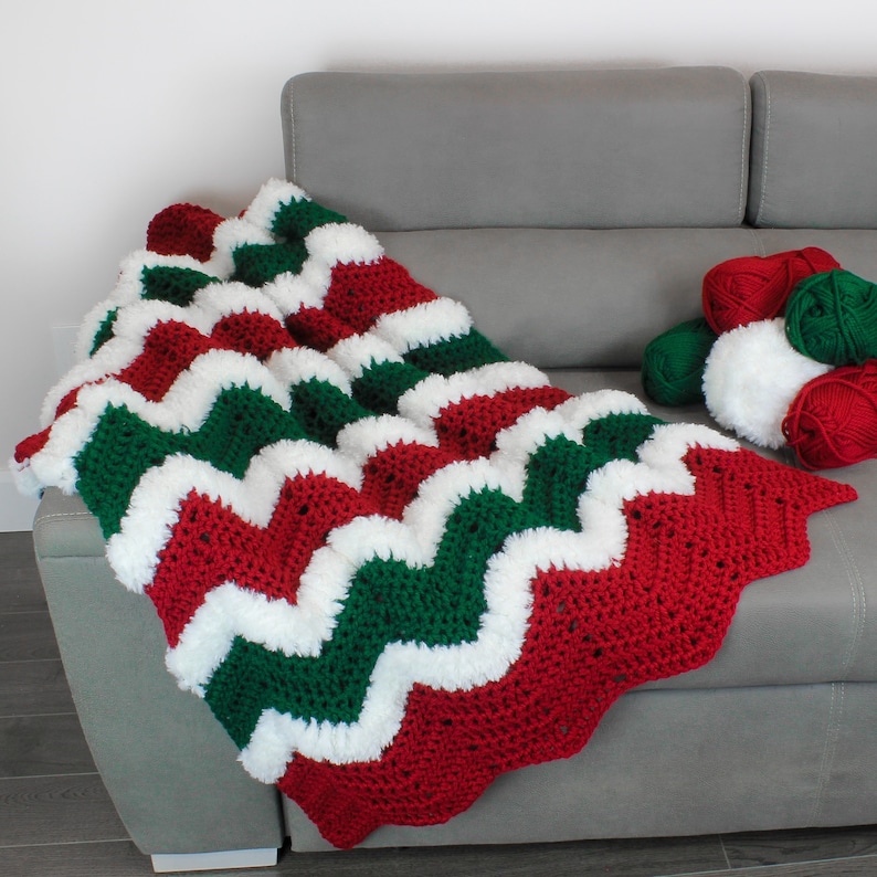 May include: A crocheted blanket with a chevron pattern in red, green, and white. The blanket is draped over a gray couch with balls of yarn in the background.