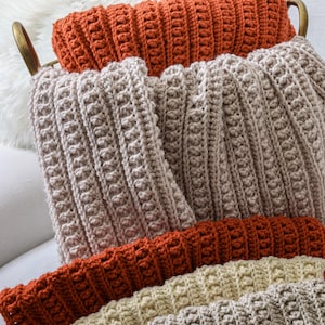 May include: A stack of crocheted throw pillows in various shades of brown, beige, and cream. The pillows are made with a textured stitch and have a soft, cozy look.