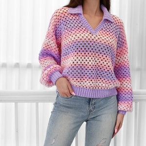 Peut inclure: Un pull en crochet ray&eacute; dans les tons de violet, rose et blanc. Le pull a un col en V et un col. Le mod&egrave;le porte le pull avec un jean bleu.