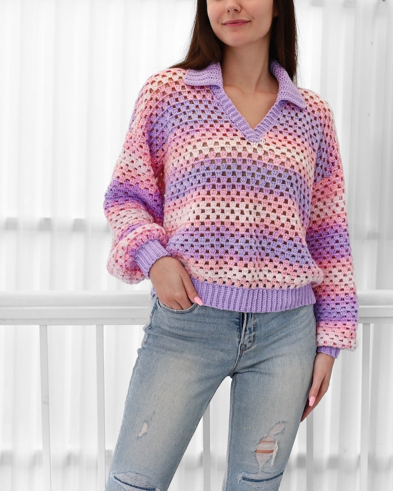 Peut inclure: Un pull en crochet ray&eacute; dans les tons de violet, rose et blanc. Le pull a un col en V et un col. Le mod&egrave;le porte le pull avec un jean bleu.
