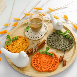May include: A set of three round crochet coasters shaped like pumpkins in orange, olive green, and cream, arranged on a round wooden tray with a glass mug of coffee, two white ceramic bird figurines, and cinnamon sticks.