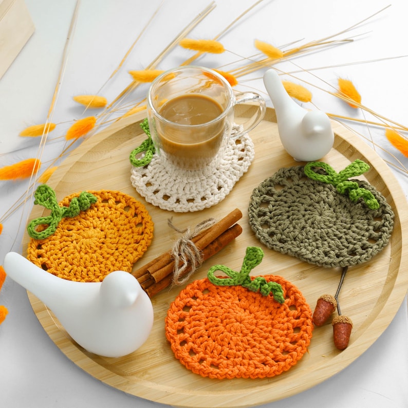 May include: A set of three round crochet coasters shaped like pumpkins in orange, olive green, and cream, arranged on a round wooden tray with a glass mug of coffee, two white ceramic bird figurines, and cinnamon sticks.