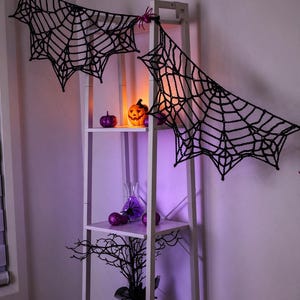 May include: A black crocheted spider web garland draped across a white ladder shelf. The shelf displays a glowing orange jack-o'-lantern, purple glittery pumpkins, a glass vase with purple spheres, and a black decorative tree branch.