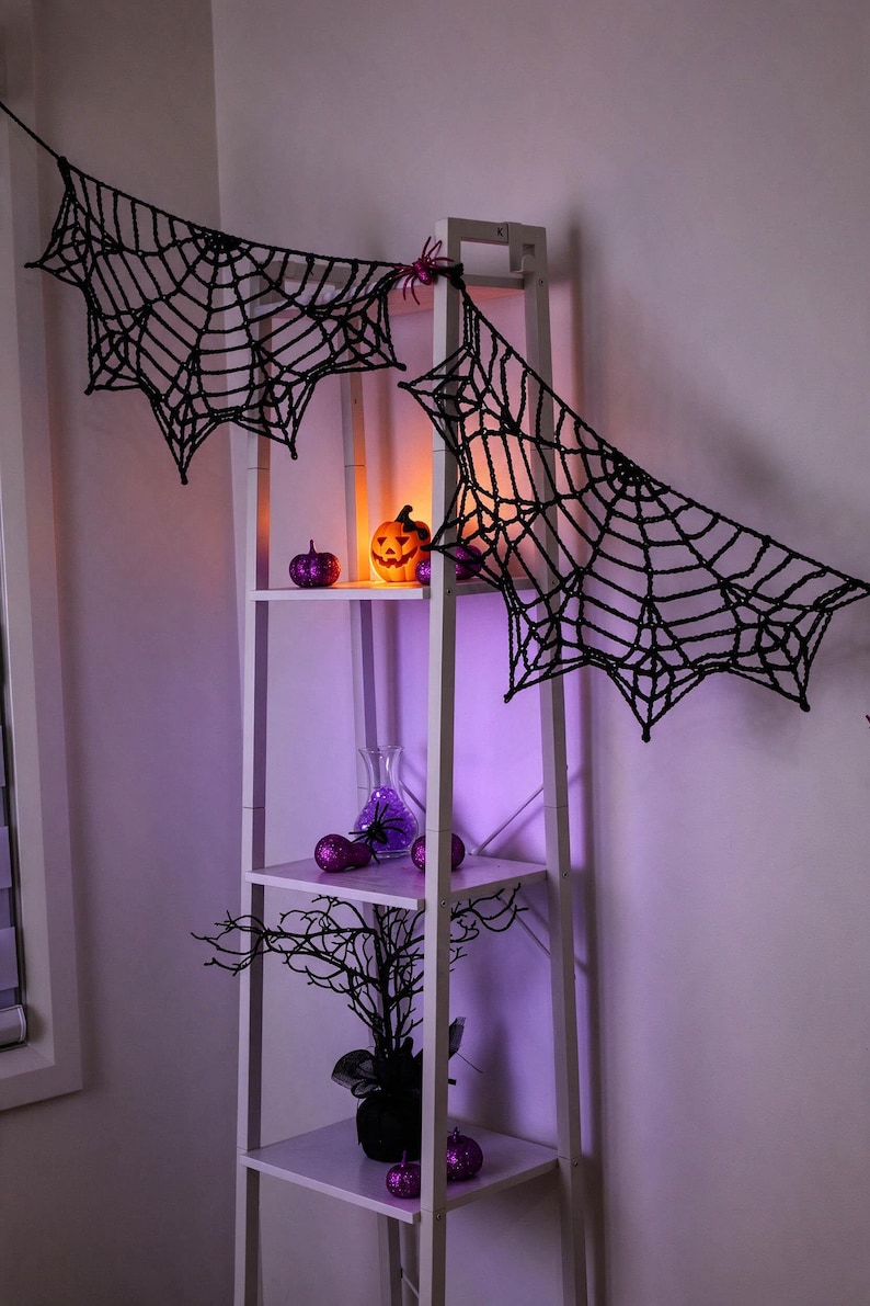 May include: A black crocheted spider web garland draped across a white ladder shelf. The shelf displays a glowing orange jack-o'-lantern, purple glittery pumpkins, a glass vase with purple spheres, and a black decorative tree branch.