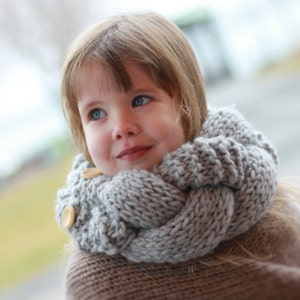 May include: A young girl wearing a gray knitted scarf with wooden buttons. The scarf is thick and cozy, perfect for keeping warm in the winter.