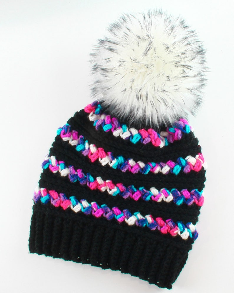 Puede incluir: Gorro de punto negro con un pomp&oacute;n de piel sint&eacute;tica blanca. El gorro tiene un patr&oacute;n de rayas de colores con hilo rosa, azul y blanco.