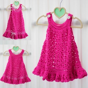 May include: A pink crocheted baby dress with a ruffled hem and white buttons. The dress is hanging on a heart-shaped hanger.