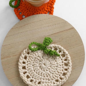 May include: A round, cream-colored crocheted coaster shaped like a pumpkin with a green stem and leaf detail. The coaster features a circular center with an openwork border. A second orange pumpkin-shaped coaster is partially visible underneath a glass.