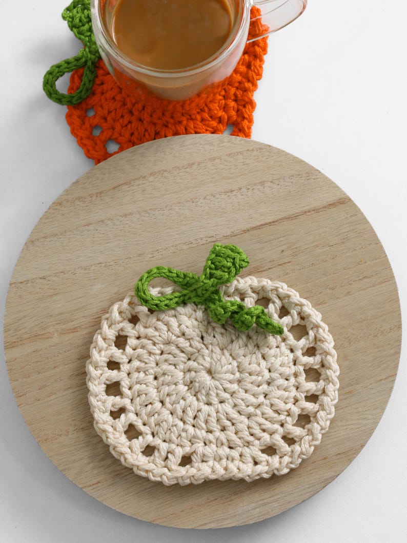 May include: A round, cream-colored crocheted coaster shaped like a pumpkin with a green stem and leaf detail. The coaster features a circular center with an openwork border. A second orange pumpkin-shaped coaster is partially visible underneath a glass.