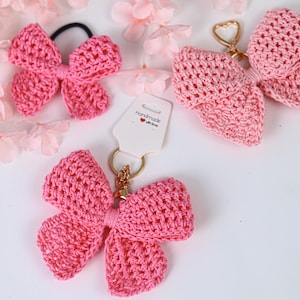 May include: Three pink crocheted bow accessories. One is a hair tie, and two are keychains with gold-toned hardware. One keychain is attached to a white display card with the text "Handmade with love" and a small red heart icon printed on it.