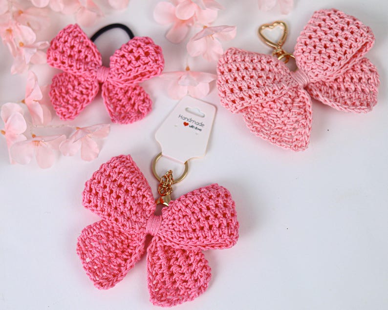 May include: Three pink crocheted bow accessories. One is a hair tie, and two are keychains with gold-toned hardware. One keychain is attached to a white display card with the text "Handmade with love" and a small red heart icon printed on it.