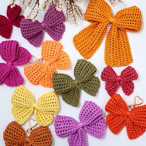 May include: A collection of crocheted bows in various colors including mustard yellow, olive green, bright orange, magenta, purple, and light yellow. Some bows feature gold-toned heart-shaped metal clasps attached to the center knot.