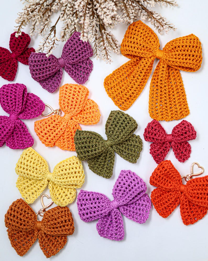 May include: A collection of crocheted bows in various colors including mustard yellow, olive green, bright orange, magenta, purple, and light yellow. Some bows feature gold-toned heart-shaped metal clasps attached to the center knot.