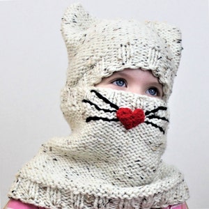 May include: A white knitted cat hat with black whiskers and a red heart nose. The hat has a hood and covers the head and neck.