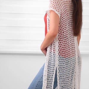 May include: A long, white, open-front crochet vest featuring a repeating hexagonal mesh pattern, short sleeves, and high side slits. The hem is finished with long, straight fringe detailing that hangs down to the lower thigh area of the garment.