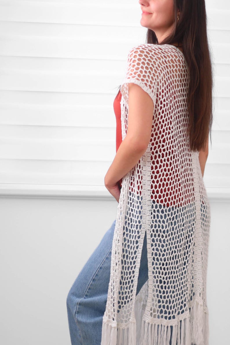 May include: A long, white, open-front crochet vest featuring a repeating hexagonal mesh pattern, short sleeves, and high side slits. The hem is finished with long, straight fringe detailing that hangs down to the lower thigh area of the garment.