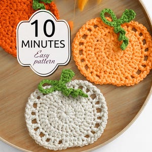 May include: Two round crochet coasters shaped like pumpkins, one orange and one off-white, each featuring a green stem and vine. A white label with a decorative border displays the text 10 MINUTES Easy pattern in black font.