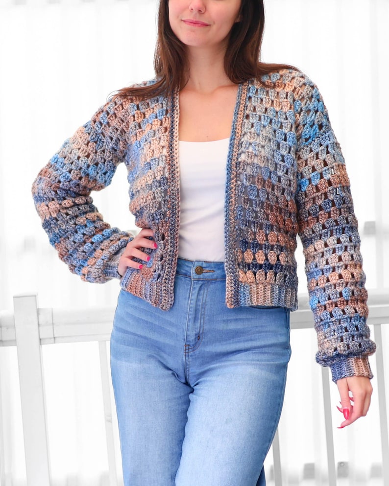 May include: A crocheted cardigan in shades of blue, tan, and brown. The open-front sweater has long sleeves and a textured, open weave design. The garment is paired with blue jeans.