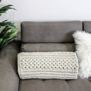 May include: A light gray crocheted throw blanket is draped over a gray couch with a white faux fur pillow.