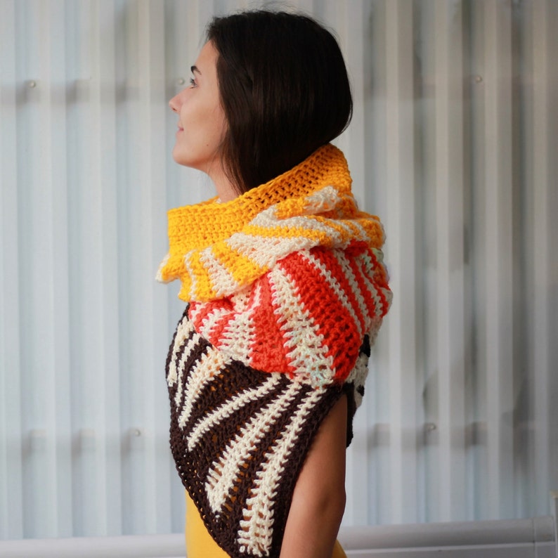 May include: A crocheted cowl in a multi-colored striped pattern. The cowl is made of brown, white, orange, and yellow yarn. The cowl is worn by a person with long brown hair.
