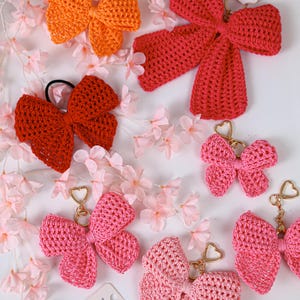 May include: A collection of crocheted bow accessories in various shades of orange, red, and pink. Several bows are attached to gold-toned heart-shaped keychains, while others feature hair ties or display tags with the text Handmade with love.