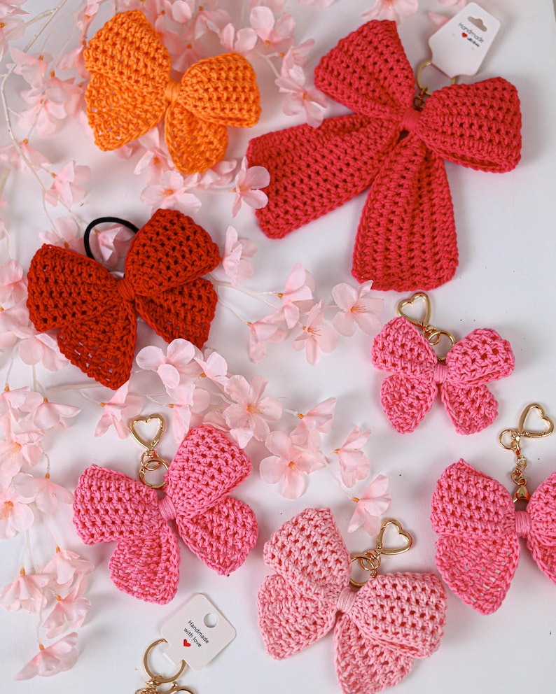 May include: A collection of crocheted bow accessories in various shades of orange, red, and pink. Several bows are attached to gold-toned heart-shaped keychains, while others feature hair ties or display tags with the text Handmade with love.