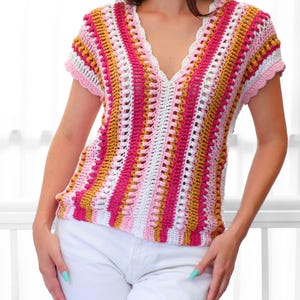 Pu&ograve; includere: Un top all'uncinetto a maniche corte e scollo a V. Il top presenta un motivo a righe nei toni del rosa, bianco e oro. I bordi sono rifiniti con un bordo smerlato. L'indumento &egrave; abbinato a pantaloni bianchi.