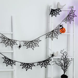 May include: A black crocheted spider web garland featuring multiple web segments connected by a string. Small black spider figures are attached to the webs. The garland is draped across a white ladder shelf, which also holds a small orange pumpkin.